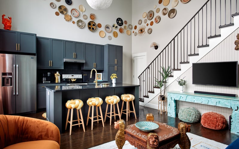 Open-plan kitchen and living room with dark blue cabinetry, wooden bar stools, decorative plates on walls, and staircase with black railing.