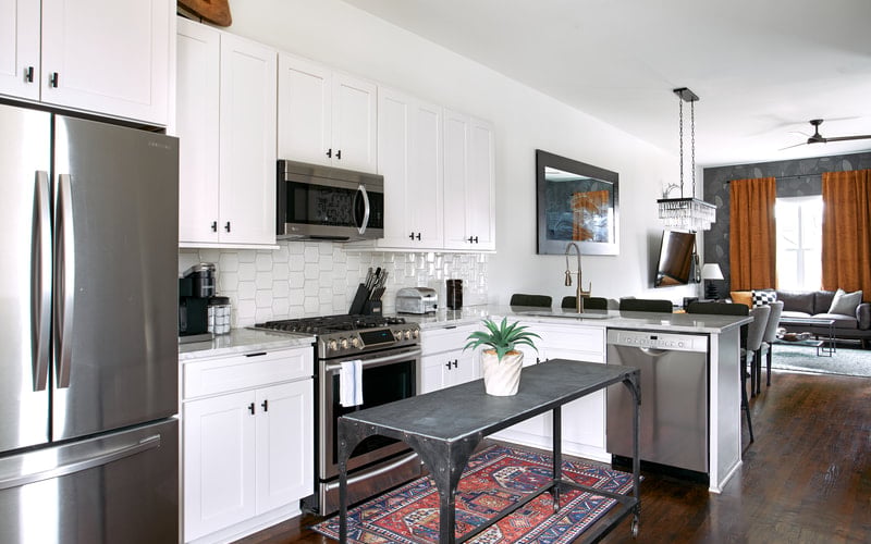 Modern kitchen with white cabinetry, stainless steel appliances, white subway tile backsplash, and black island with potted plant, open to living room with