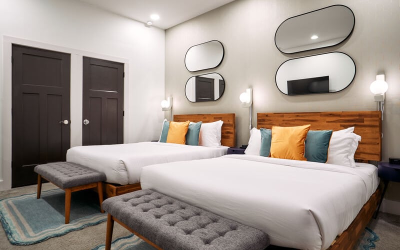 Modern bedroom with two queen beds, wooden headboards, decorative mirrors, dark doors, and contemporary lighting fixtures