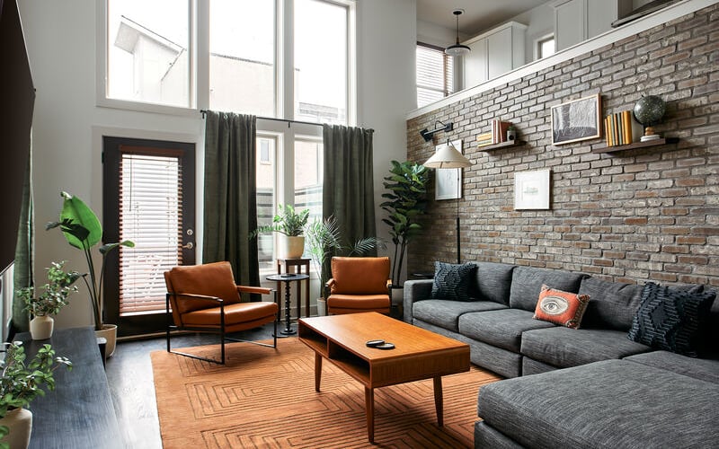 Modern living room with gray sectional sofa, orange mid-century chairs, exposed brick wall, wood coffee table, and potted plants