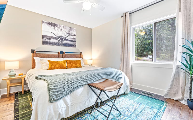 Bright bedroom with wood bed frame, white bedding, blue striped throw, large windows overlooking trees, and ceiling fan