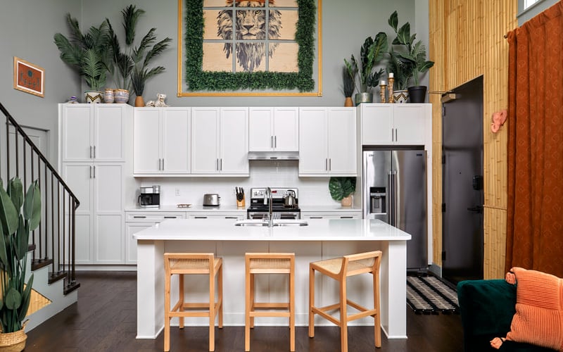 Modern kitchen with white cabinetry, stainless steel appliances, island with wooden bar stools, and potted plants throughout