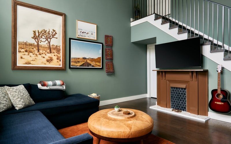Modern living room with sage green walls, navy sectional sofa, round wooden ottoman, fireplace with TV, and floating staircase with metal railing