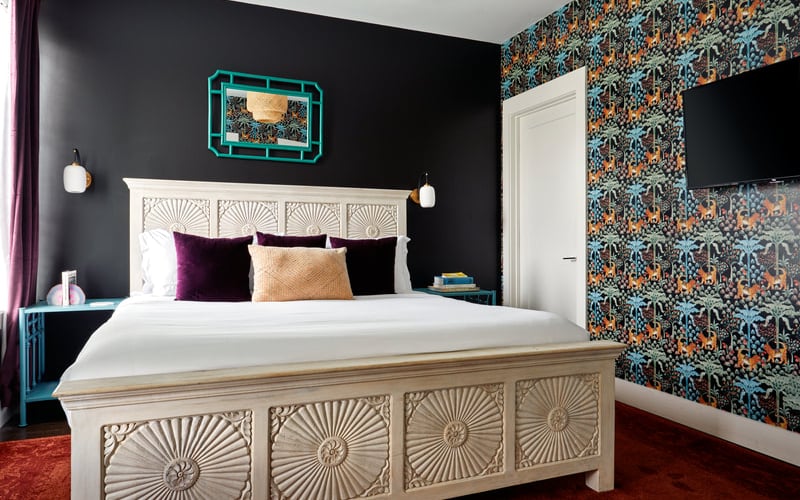 Bedroom with carved wooden bed, black walls, neon turquoise artwork, patterned wallpaper accent wall, and burgundy rug