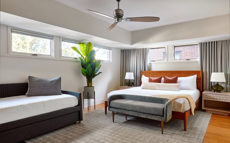 Bright bedroom with two beds, wood furniture, potted plant, ceiling fan, and multiple windows with natural light