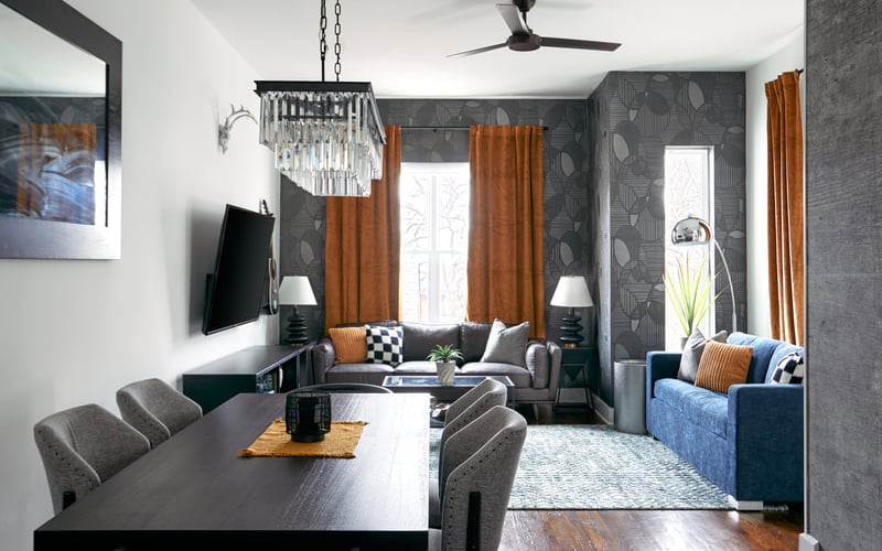 Open-plan living and dining area with gray patterned walls, orange curtains, dark furniture, and crystal chandelier