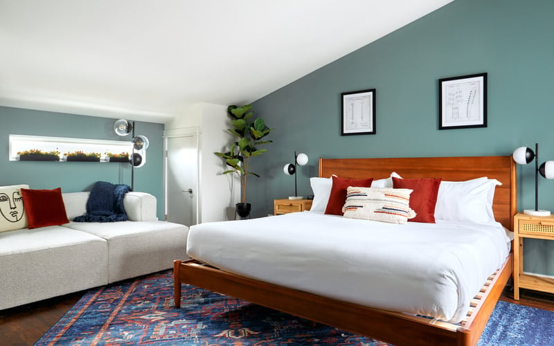 Bedroom with teal walls, wooden bed with white bedding and red pillows, cream sofa, blue patterned rug, and framed artwork