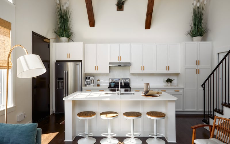 Modern white kitchen with island, stainless steel appliances, four bar stools, and exposed wood beams