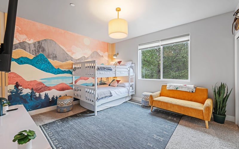 Bright children's bedroom with bunk beds, colorful mountain mural, orange sofa, and large windows overlooking trees