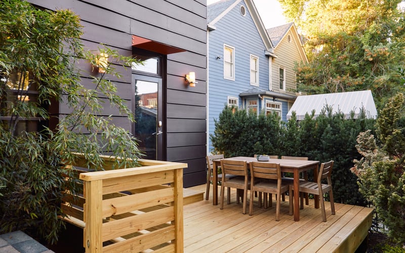 Wooden deck with dining table and chairs, bordered by dark modern house and blue Victorian home, surrounded by greenery
