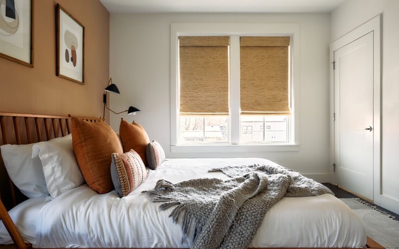Bedroom with wood-frame bed, white bedding, orange and patterned pillows, woven window shades, and tan accent wall.