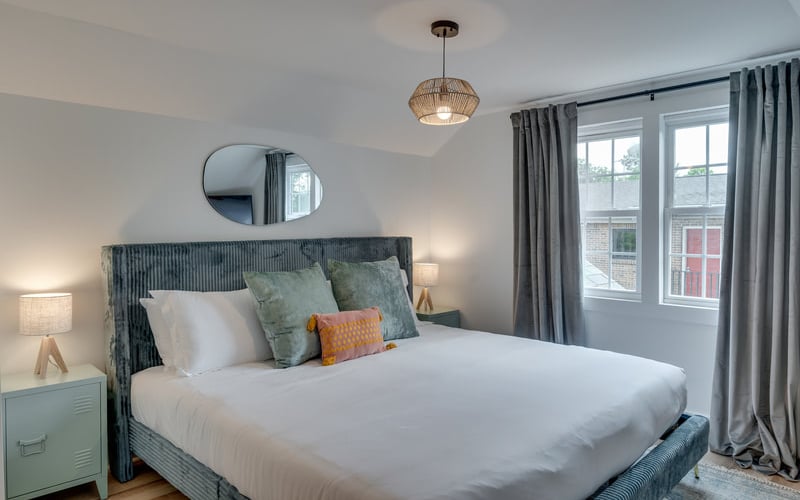 Bright bedroom with textured gray upholstered bed, white bedding, decorative pillows, pendant light, round mirror, and gray curtains framing windows with