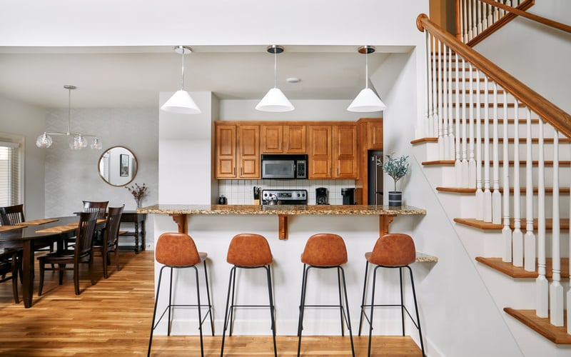 Open-plan kitchen and dining area with wooden cabinets, granite countertop, four orange bar stools, and wooden staircase with white railings.