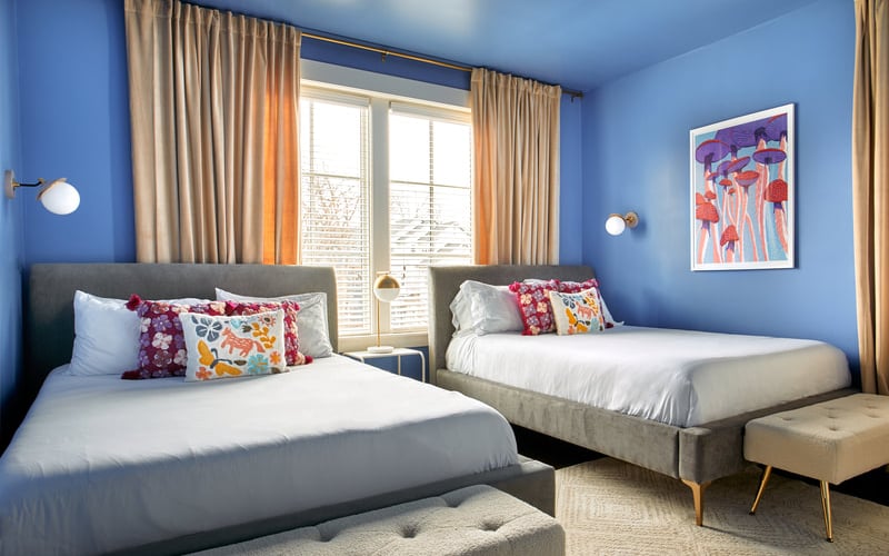 Bright blue bedroom with two gray beds, colorful throw pillows, beige curtains, and framed mushroom artwork
