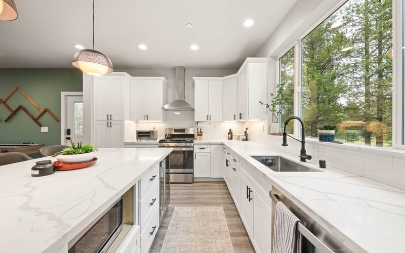 Modern kitchen with white cabinetry, marble countertops, stainless steel appliances, and large windows overlooking trees