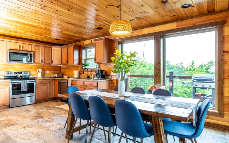 Wood-paneled kitchen and dining area with blue chairs, large windows overlooking green landscape, and pendant lighting