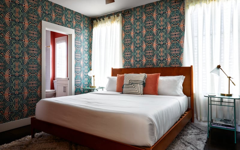 Bedroom with patterned teal and coral wallpaper, wooden bed frame, white bedding, and sheer curtains by windows