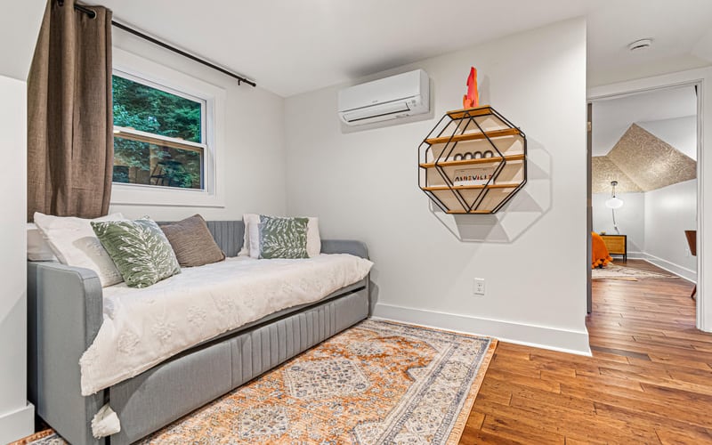 Bright bedroom with gray daybed, patterned pillows, wooden floor, geometric wall art, and view into adjoining room