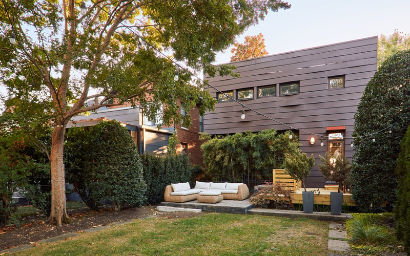 Modern dark-clad house with backyard patio, wicker seating, mature trees, and ivy-covered walls