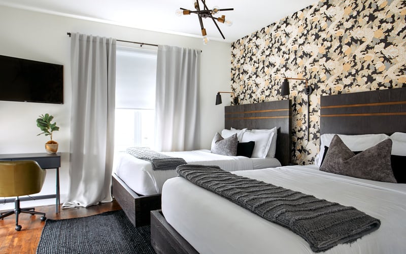 Modern hotel bedroom with two queen beds, patterned accent wall, desk with chair, and window with white curtains