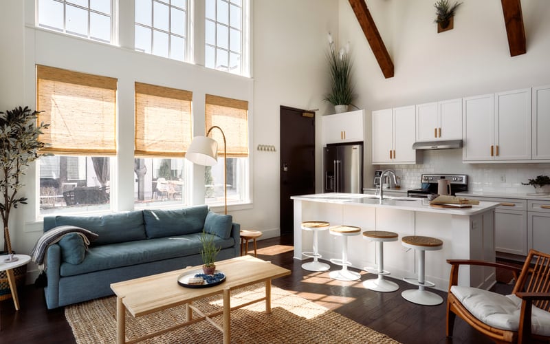 Open-plan kitchen and living room with blue sofa, white cabinetry, island seating, and large windows with natural light