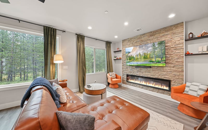 Modern living room with cognac leather sofa, brick fireplace, forest views through large windows, and orange accent chairs