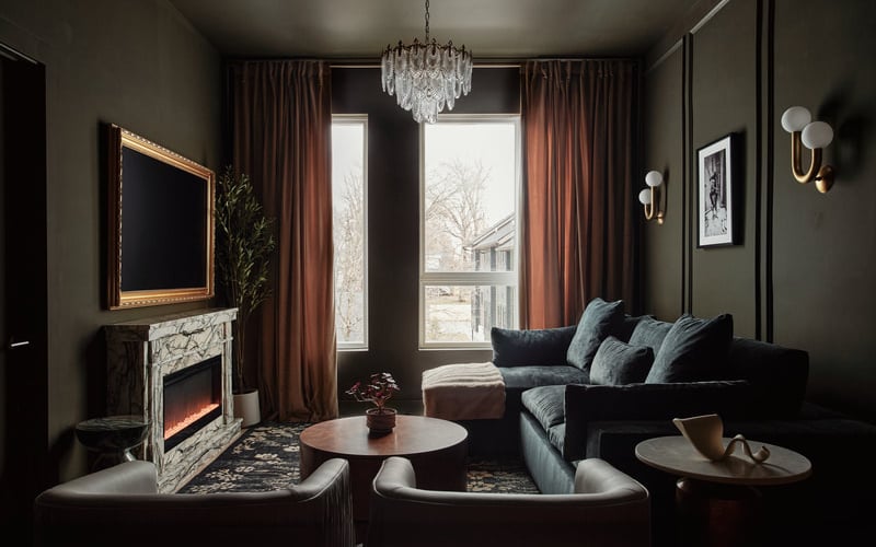 Elegant living room with dark walls, marble fireplace, black sofa, crystal chandelier, and terracotta curtains framing tall windows.