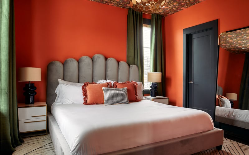Bedroom with bright orange walls, gray upholstered headboard, white bedding, and black door