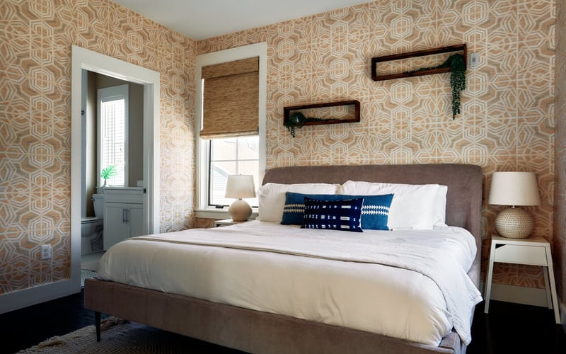 Bedroom with patterned tan wallpaper, gray upholstered bed, white bedding, blue accent pillow, and floating black shelves above headboard