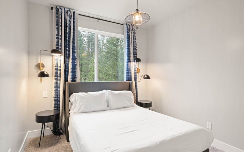 Modern bedroom with four-poster bed, white bedding, blue patterned curtains, pendant lights, and forest view through large window
