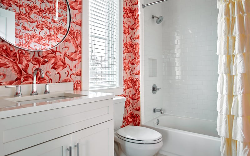 Bathroom with red patterned wallpaper, white vanity, round mirror, toilet, and bathtub with shower and yellow towel rack