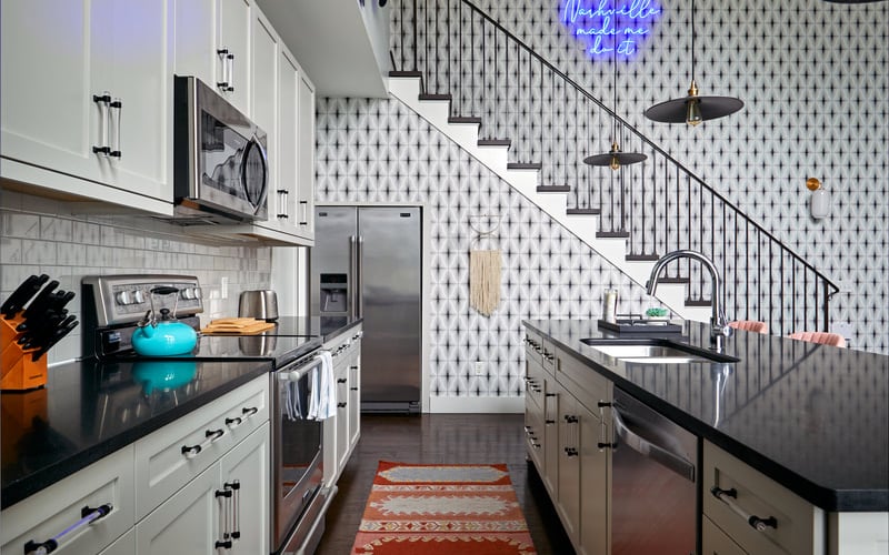Modern kitchen with white and gray cabinetry, black countertops, stainless steel appliances, and stairs with metal railing visible in background
