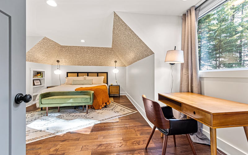 Modern bedroom with textured ceiling feature, wood bed frame, green bench, desk, and forest view through large window