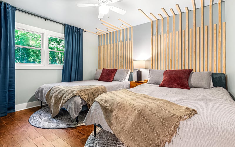 Bright bedroom with two queen beds, wooden slat headboard, blue curtains, ceiling fan, and forest view from window