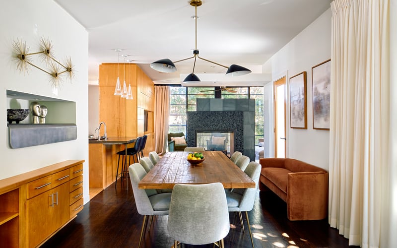 Open-plan dining and kitchen area with wood table, upholstered chairs, orange cabinetry, and dark fireplace.