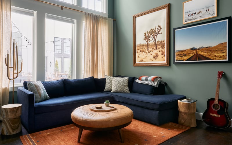 Living room with navy sectional sofa, round wooden ottoman, desert landscape artwork, and red acoustic guitar against sage green walls