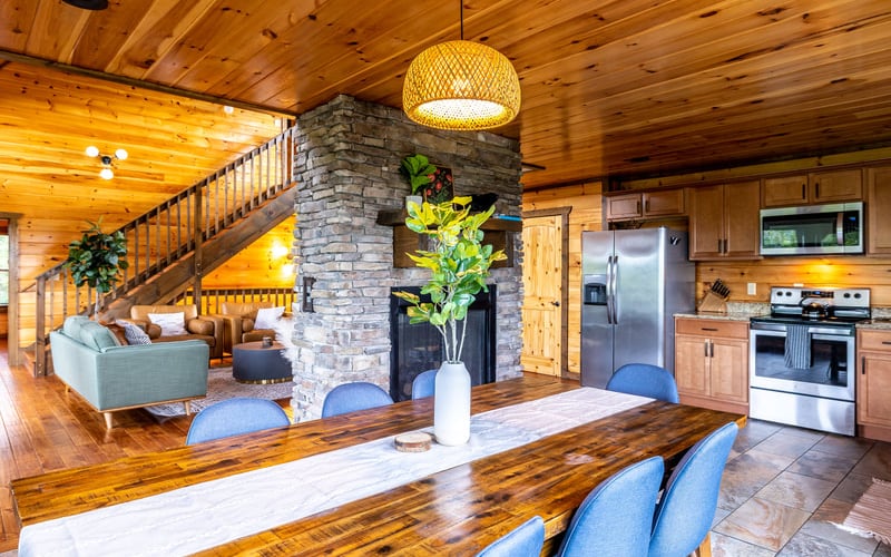 Open-plan cabin interior with wood ceilings, dining table, stone fireplace, and kitchen with stainless steel appliances