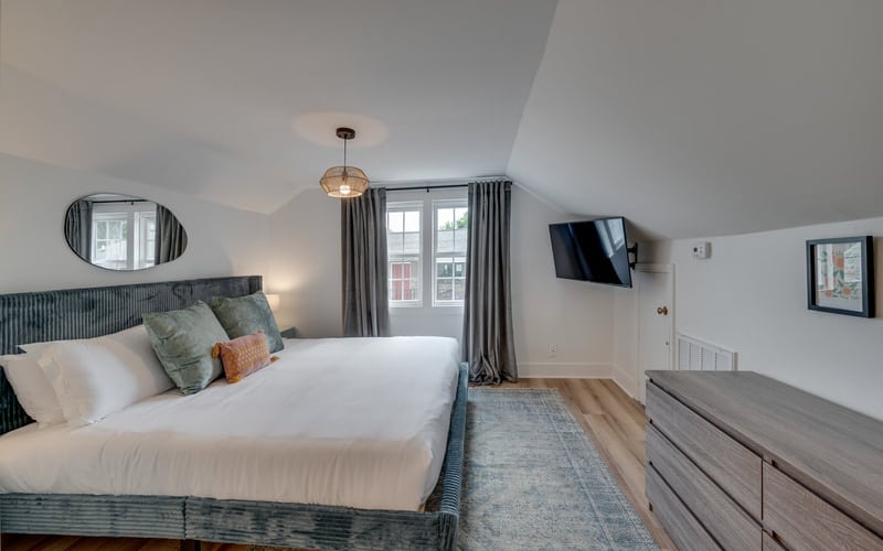 Bright attic bedroom with queen bed, gray headboard, wall-mounted TV, dresser, and doors opening to balcony