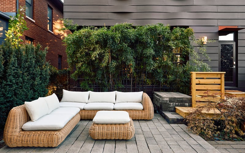 Modern courtyard patio with wicker sectional sofa, white cushions, and wooden storage box, surrounded by bamboo and brick buildings.