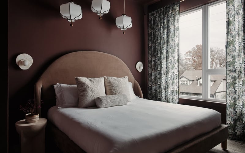 Modern bedroom with burgundy walls, curved upholstered headboard, white bedding, pendant lights, and large windows overlooking suburban homes.