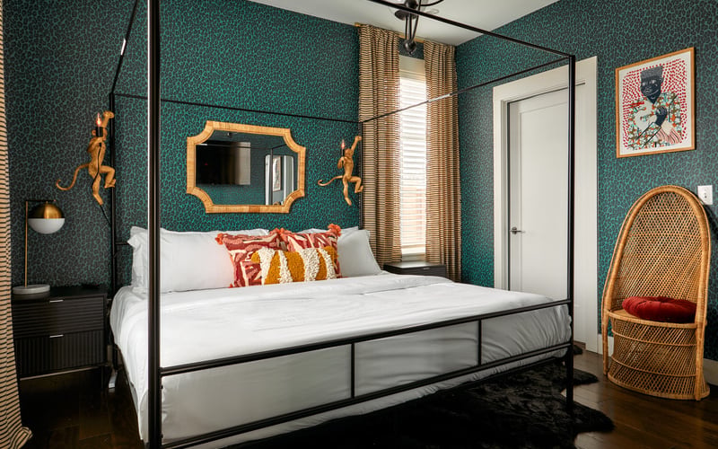Bedroom with teal patterned wallpaper, black four-poster bed, gold-framed mirror, woven chair, and gold curtains