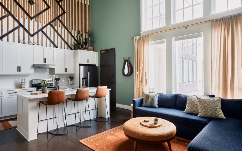 Open-plan kitchen and living room with white cabinetry, sage green walls, navy sectional sofa, and wooden staircase with geometric railing.