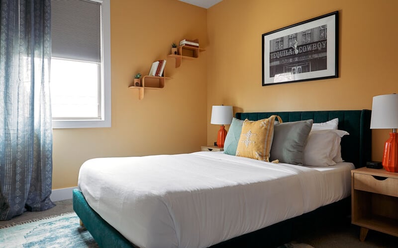 Bedroom with mustard-yellow walls, dark teal bed, white bedding, wooden nightstands with orange lamps, and window with blue curtains