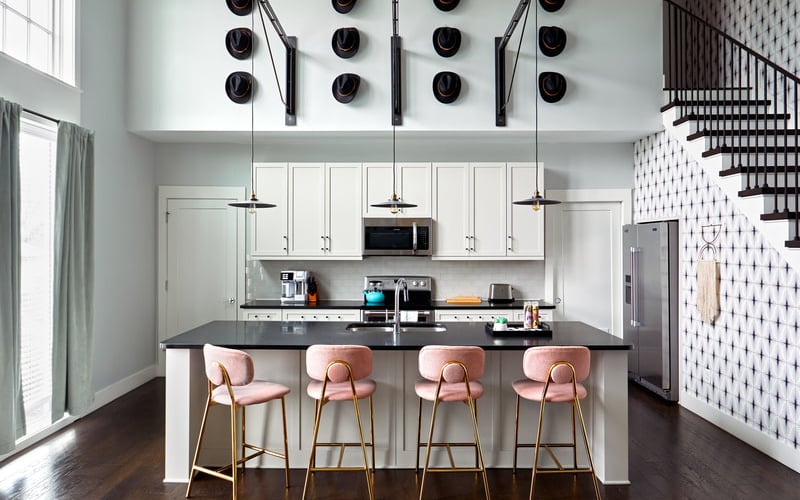 Modern kitchen with white cabinetry, black countertop island, four pink bar stools, pendant lights, and decorative black hats on wall