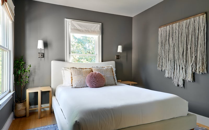 Bedroom with gray walls, white bed, patterned pillows, macramé wall hanging, and window with garden view