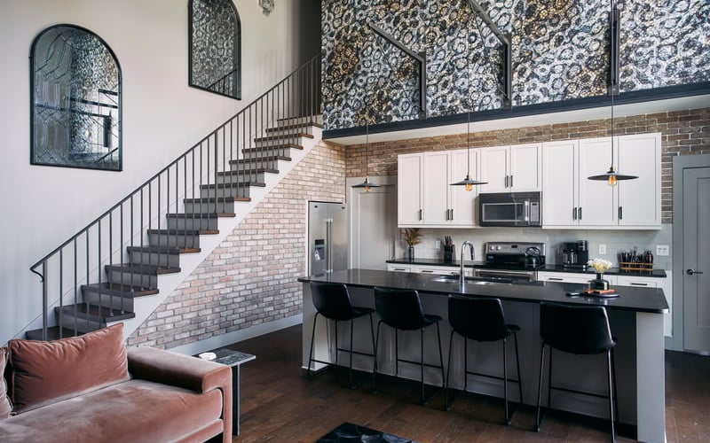 Modern loft kitchen with white cabinetry, black island, brick staircase, and patterned wall panels above