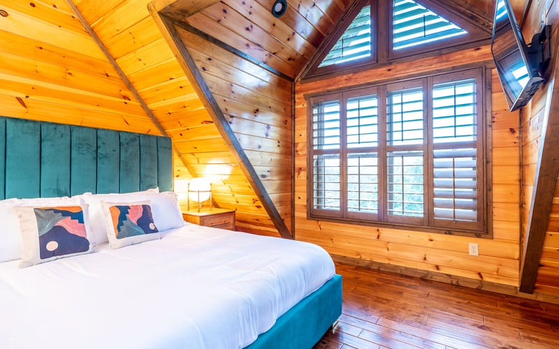 Wooden cabin bedroom with vaulted ceiling, queen bed with teal headboard, and large shuttered windows
