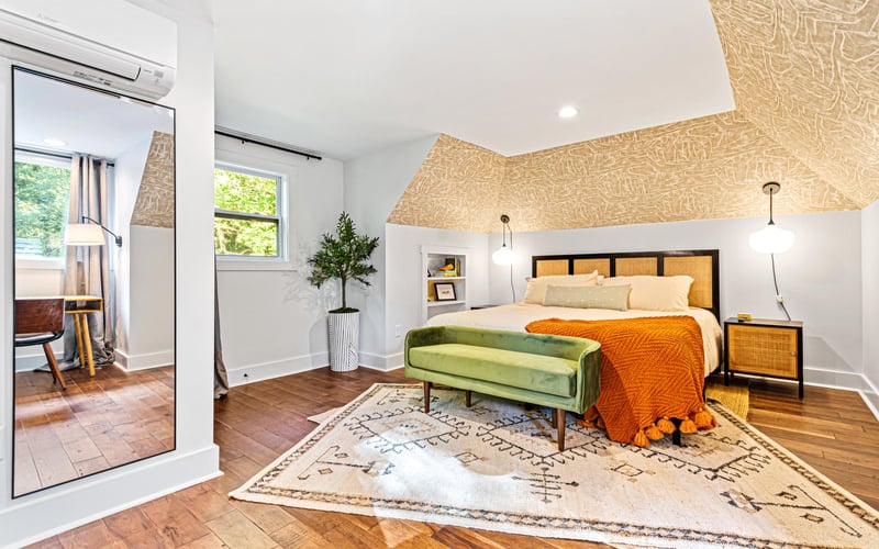 Bright attic bedroom with sloped textured ceiling, wood bed frame, green bench, orange throw, and hardwood floors.