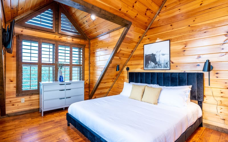 Wooden cabin bedroom with vaulted ceiling, queen bed with dark headboard, white dresser, and glass doors opening to outdoor view.