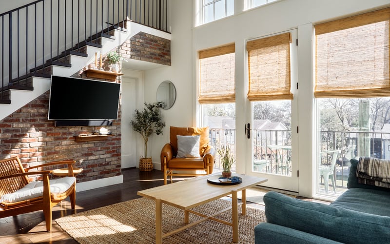 Modern living room with exposed brick wall, wooden furniture, TV, and glass doors opening to a balcony with winter trees visible outside.
